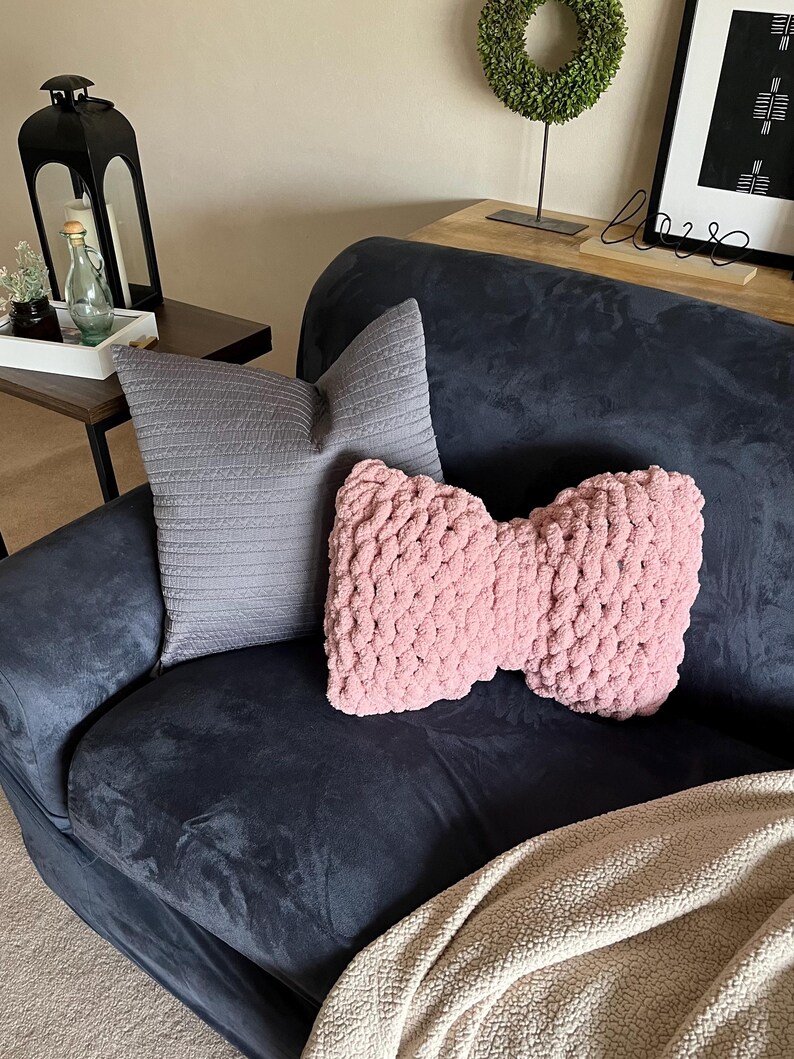 May include: A navy blue sofa with two decorative pillows and a blanket. One pillow is a light gray, rectangular shape. The other is a pink, bow-shaped, chunky knit pillow. A beige blanket is draped over the sofa.