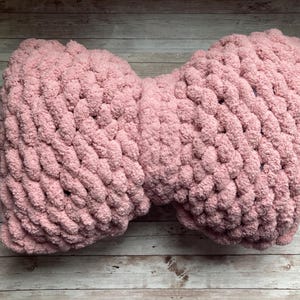 May include: A soft, pink, chunky-knit pillow in a bow shape. The pillow is made of a thick, plush yarn and is resting on a light-colored wooden surface. The texture of the yarn is visible.