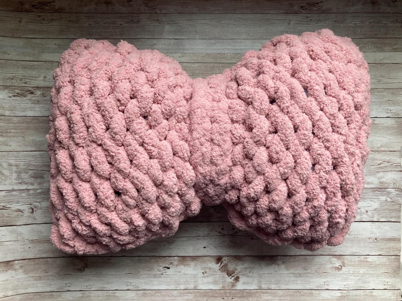 May include: A soft, pink, chunky-knit pillow in a bow shape. The pillow is made of a thick, plush yarn and is resting on a light-colored wooden surface. The texture of the yarn is visible.