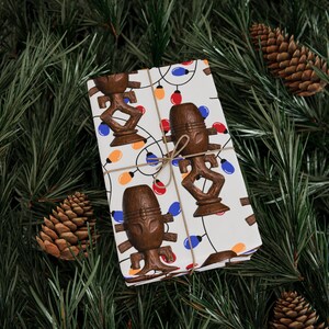 May include: A wrapped gift with a festive design featuring brown tiki figures and colourful string lights on a white background. The gift is tied with twine and set against a backdrop of pine needles and pine cones.
