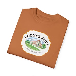Może przedstawiać: Brązowa koszulka z białym logo z napisem "Boone's Farm American Original" i kreskówkowym obrazkiem wiejskiego domu.