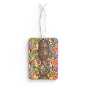 May include: A rectangular air freshener with a brown wooden tiki statue design. The background is a colourful floral pattern with various fruits and flowers. A white string is attached for hanging.
