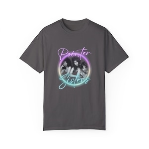 May include: Dark gray t-shirt featuring a black and white image of the Pointer Sisters inside a circular design with the words "Pointer Sisters" in a neon-style font. The t-shirt is made of a soft material.
