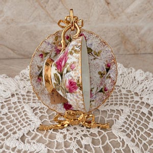 May include: A porcelain teacup and saucer set featuring a pink rose floral pattern with gold trim, displayed on a gold-toned metal stand. The teacup has a gold handle and the saucer has a scalloped edge with a speckled gold finish.