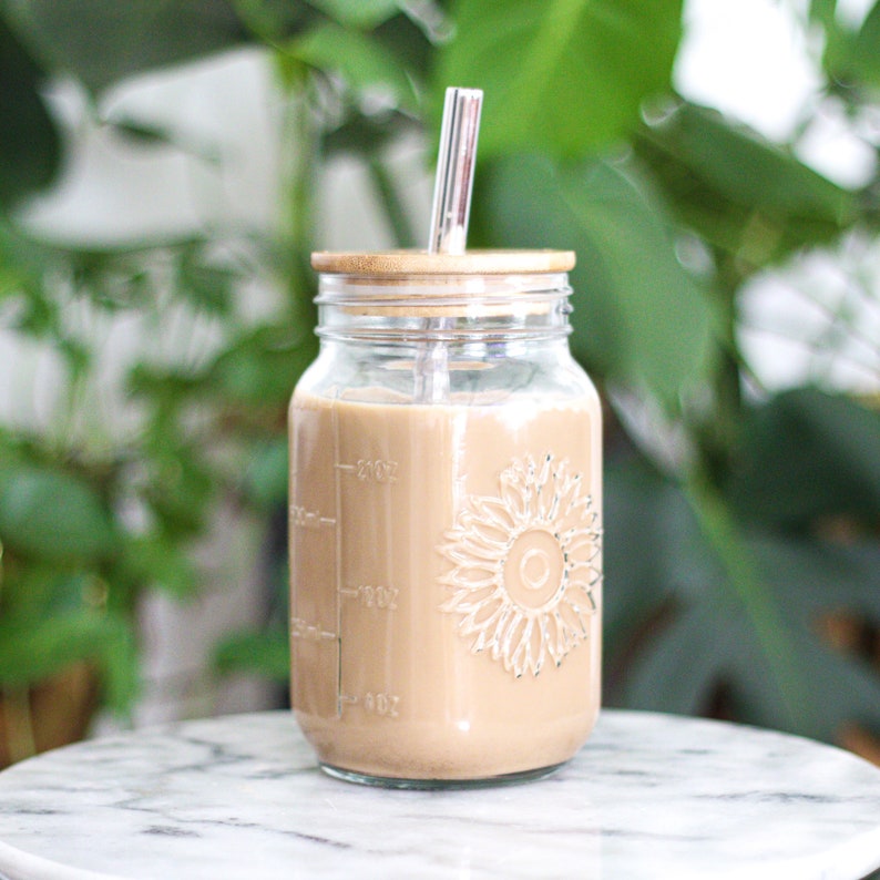 May include: A clear glass mason jar with a sunflower design, filled with a light brown beverage. It has a bamboo lid and a clear glass straw. The jar has measurement markings on the side. The background is blurred green foliage.