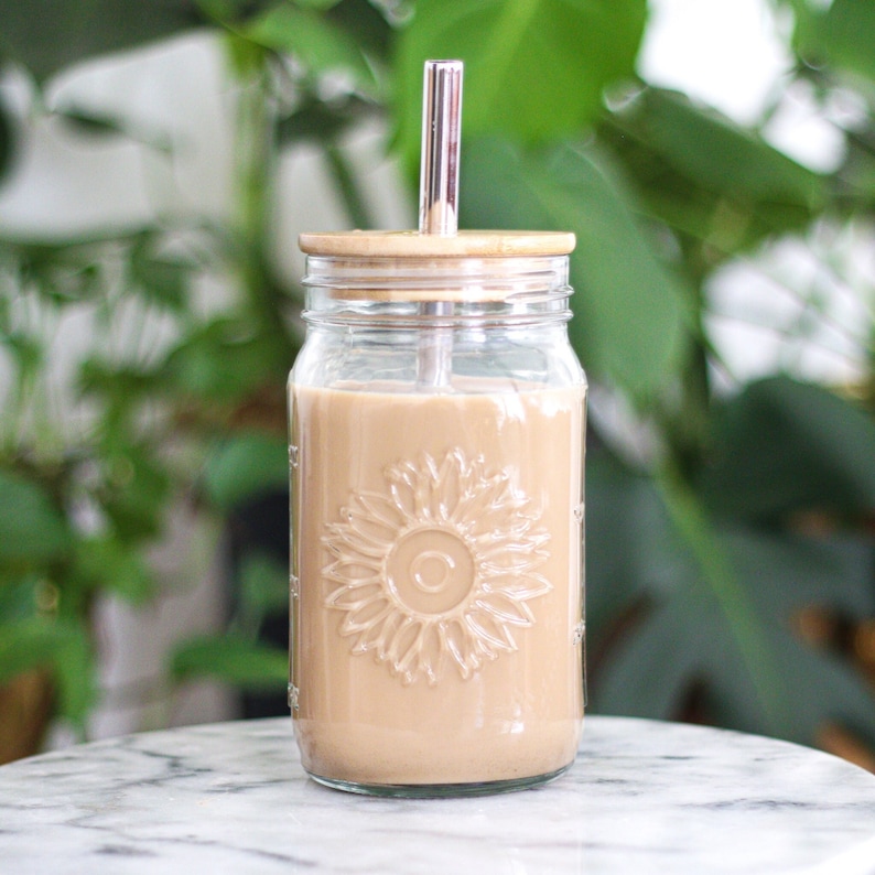 May include: A clear glass mason jar with a sunflower design filled with a light brown beverage. It has a bamboo lid and a silver metal straw. The jar is sitting on a white marble surface.