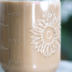 May include: A clear glass jar filled with a light brown liquid, possibly coffee or tea. The jar has embossed sunflower detailing and measurement markings in ounces. The liquid fills the jar to the 20 oz mark.