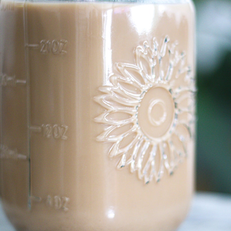 May include: A clear glass jar filled with a light brown liquid, possibly coffee or tea. The jar has embossed sunflower detailing and measurement markings in ounces. The liquid fills the jar to the 20 oz mark.