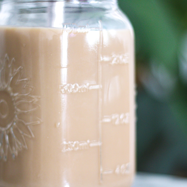 May include: A clear glass jar filled with a light brown liquid, possibly a beverage. The jar has measurement markings in milliliters and a sunflower design on the side. The background is a blurred green.