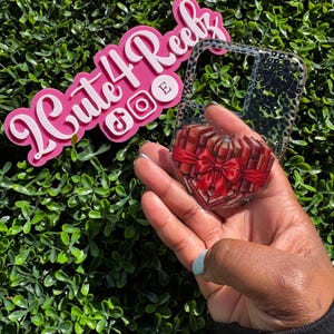May include: Clear phone case with a heart-shaped design of books tied with a red bow. The case is held in front of a green leafy background. A pink sign with the text "Cute4Reels" and social media icons is in the background.