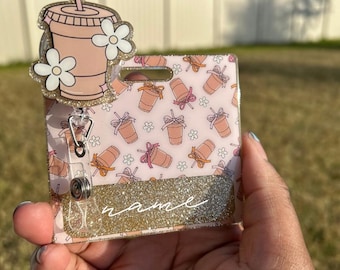 May include: A pink and gold glitter ID badge holder with a cartoon image of a pink iced coffee with a straw and white flowers. The holder has a clear plastic retractable badge reel and a pink and white patterned background with a space for a name.
