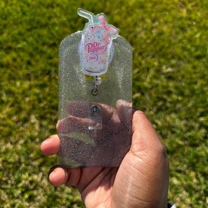 May include: A clear glitter badge holder with a pink and white Dr Pepper Diet can charm attached. The charm has a straw and a pink bow.