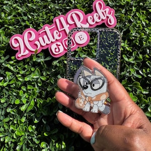 May include: A clear phone case with a cartoon dog character design. The dog is wearing glasses and a scarf. The image also includes a pink sign that says "Cute 4 Reebz".