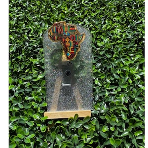 May include: A badge reel with a glittery, clear resin design. The badge reel features a colorful map of Africa with gold trim. The reel is attached to a wooden stand, set against a backdrop of green foliage.