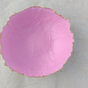 May include: A handmade, round pink bowl with a textured surface and a gold-colored, uneven edge. The bowl is centered on a white, textured fabric background. This decorative bowl is ideal for serving snacks or as a decorative piece.