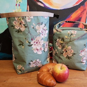 Könnte beinhalten: Zwei Stoff-Lunch-Taschen mit floralem Muster. Die Taschen sind hellgrün mit weißen und rosa Kirschblüten. Eine Tasche ist hoch mit einem beigen Riemen und Schnalle. Die andere Tasche ist kürzer mit einem Griff. Ein Croissant und ein Apfel liegen vor den Taschen.