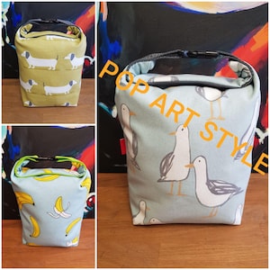 Könnte beinhalten: Drei Stoff-Lunchbags mit verschiedenen Mustern. Eine Tasche ist hellgrün mit Dackeln, eine andere hellblau mit Möwen und die dritte hellblau mit Bananen. Jede Tasche hat einen Griff und einen schwarzen Schnallenverschluss.
