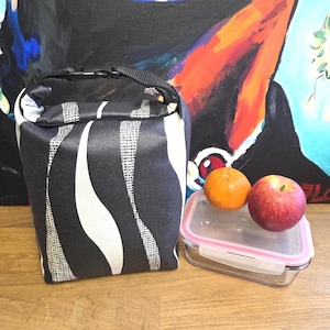 Könnte beinhalten: Eine schwarz-weiß gemusterte Lunchtasche mit schwarzem Tragegriff und Schnallenverschluss, neben einem rechteckigen Vorratsbehälter aus klarem Glas mit rosa Deckel, einem roten Apfel und einer kleinen Orange.