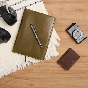 May include: An overhead shot of a collection of items on a wooden surface. Includes a dark green leather journal, a black pen with copper accents, a black camera, a brown leather wallet, and black headphones.