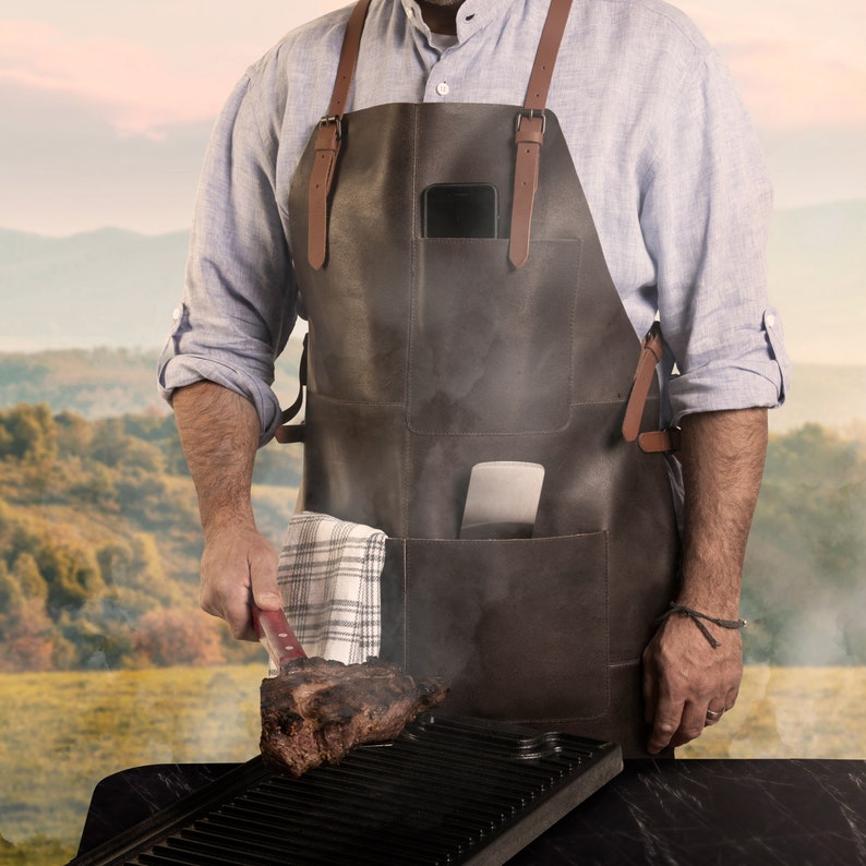 May include: A dark brown leather apron with brown leather straps and pockets. The apron has a pocket holding a smartphone and a spatula. A checkered towel hangs from a pocket. A large steak is on a grill.