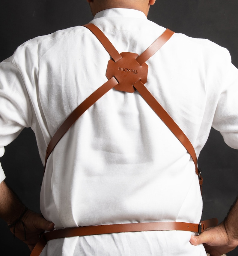 May include: Brown leather apron harness with the text "THEODORE" embossed on the back piece. The harness features adjustable straps and a cross-back design. The apron is worn over a white shirt.