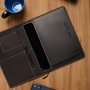 Peut inclure: Un journal en cuir noir ouvert avec le texte "THEODORE Leather" et "Journal" imprimé sur la couverture. Le journal est ouvert, révélant une tablette et un stylo. Une tasse bleue et un smartphone sont également visibles.