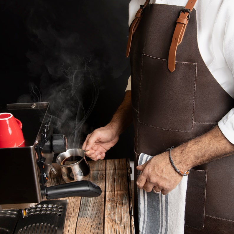 May include: A barista pouring coffee from a small stainless steel pot. The coffee machine is black with a red cup on top. The barista wears a brown leather apron with tan straps. Steam rises from the coffee, and the background is black.