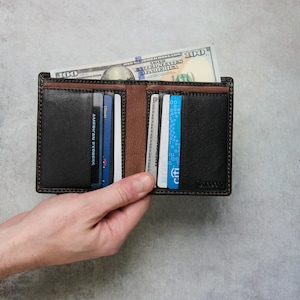Può includere: Un portafoglio a due ante in pelle nera e marrone, pieno di carte di credito e una banconota da cento dollari. Il portafoglio è aperto, mostrando gli scomparti per carte e il vano contanti. Il portafoglio ha un aspetto strutturato.