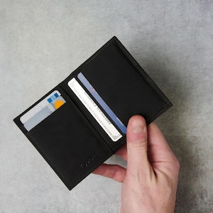 Peut inclure: Portefeuille en cuir noir ouvert, révélant des cartes de crédit. Le portefeuille présente plusieurs fentes pour cartes et un design minimaliste. Le nom de la marque "BLACK OAK" est subtilement estampé en bas. Un main tient le portefeuille ouvert.