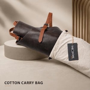 May include: A rolled dark brown leather apron with tan leather straps and a metal buckle. The apron is partially inside a cream-colored cotton carry bag. A black tag with the word "THEODORE" is attached to the bag. The words "COTTON CARRY BAG" are at the bottom.