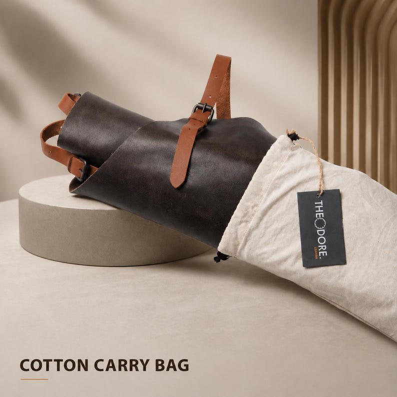 May include: A rolled dark brown leather apron with tan leather straps and a metal buckle. The apron is partially inside a cream-colored cotton carry bag. A black tag with the word "THEODORE" is attached to the bag. The words "COTTON CARRY BAG" are at the bottom.