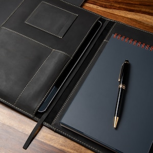 Peut inclure: Portefeuille en cuir noir avec surpiqûres crème, contenant une tablette et un carnet à spirales bleu foncé. Un stylo noir et doré repose sur le carnet. Le portefeuille a des poches et un signet, sur une surface en bois.