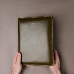 May include: An olive green leather notebook or journal. The cover has a textured finish and is held by two hands. The edges are stitched with a matching thread. The background is a plain, neutral colour.