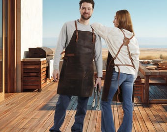 Puede incluir: Dos personas con delantales de cuero marrón con correas de cuero, de pie en una terraza de madera. Los delantales parecen estar diseñados para asar a la parrilla o cocinar. Una persona sostiene unas pinzas para asar. Una parrilla y una mesa de comedor al aire libre están en el fondo.