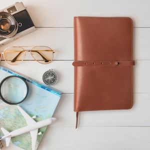May include: A brown leather journal with a strap closure, a vintage camera, sunglasses, a compass, a magnifying glass, a map, and a toy airplane are arranged on a white wooden surface. Travel and adventure theme.