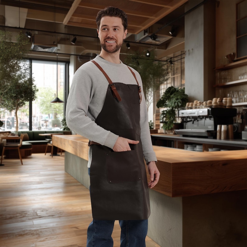 May include: A dark brown leather apron with brown leather straps and a large front pocket. The apron is worn over a gray long-sleeve shirt and blue jeans. The background shows a cafe setting with wooden tables and a coffee machine.