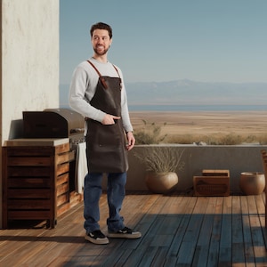 May include: A brown leather apron with brown leather straps. The apron has a large front pocket and is worn by a person. The person is wearing a gray long-sleeve shirt and blue jeans. The background includes a grill and a scenic view of a desert landscape.