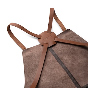 May include: Close-up of a brown leather apron with a unique design. The apron features a hexagonal leather piece at the top with the word "THEODORE" embossed on it. Brown leather straps extend from the top piece and attach to the apron.