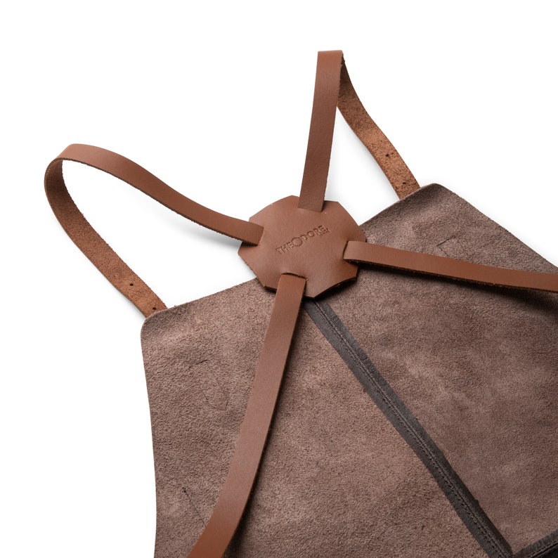May include: Close-up of a brown leather apron with a unique design. The apron features a hexagonal leather piece at the top with the word "THEODORE" embossed on it. Brown leather straps extend from the top piece and attach to the apron.