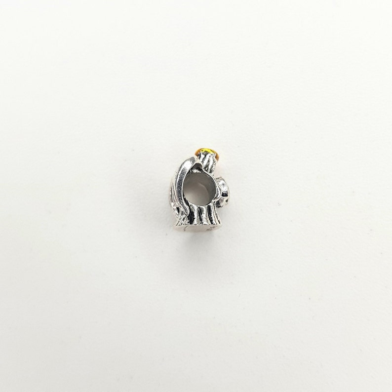 May include: Silver charm in the shape of an angel with a yellow halo. The angel is holding its hands together in prayer.