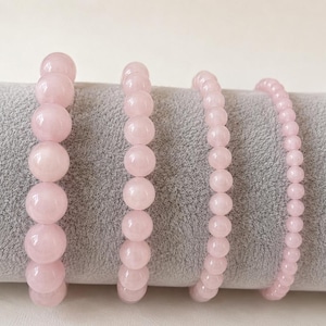 Può includere: Vengono presentati quattro braccialetti di perline di quarzo rosa di varie dimensioni. I braccialetti sono di un rosa tenue e hanno una finitura liscia e lucida. Le perline variano di dimensione, creando un effetto a strati.
