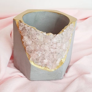 May include: A hexagonal concrete planter with a gold rim and a pink quartz crystal design. The planter is sitting on a pink fabric surface.