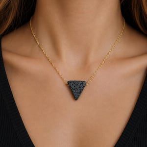 May include: A necklace featuring a triangular pendant made of black porous lava stone suspended from a thin gold-toned metal chain. The pendant has a textured surface with small holes, and the chain connects to the top corners of the triangle.