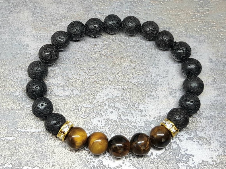 Puede incluir: Un brazalete de cuentas de lava negra y ojo de tigre con separadores dorados. El brazalete tiene un cord&oacute;n el&aacute;stico extensible.