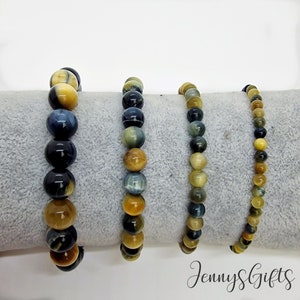 May include: Four beaded bracelets with tiger's eye stones in varying sizes. The beads display a mix of golden yellow, brown, and dark blue hues. The bracelets are displayed on a gray surface, with the text "JennyS Gifts" in the bottom right corner.