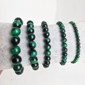 May include: Four green gemstone bracelets with round beads. The bracelets are arranged in a row on a grey surface.