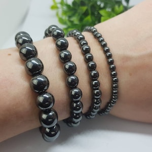 May include: Four dark grey beaded bracelets of varying sizes are displayed. The bracelets are made of smooth, round beads. The beads have a shiny, reflective surface. The bracelets are worn on a wrist.