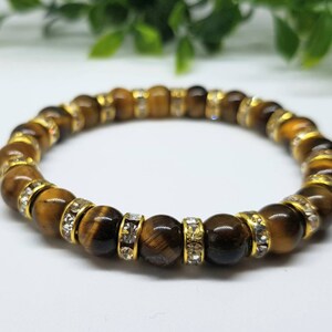 May include: A brown tiger's eye gemstone bracelet with gold accents and clear rhinestones.