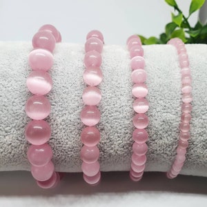 Puede incluir: Cuatro pulseras de piedras preciosas rosas con un acabado liso y pulido. Las pulseras están hechas de cuentas redondas y están dispuestas en fila sobre una superficie suave y gris.