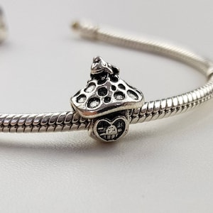 Könnte beinhalten: Silberarmband mit einem Pilz-Anhänger, auf dem sich ein Frosch befindet. Der Anhänger hat eine herzförmige Basis.